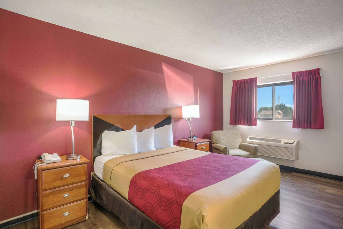 Habitación de hotel con una cama grande, dos mesitas de noche con lámparas, una silla y una ventana con cortinas.