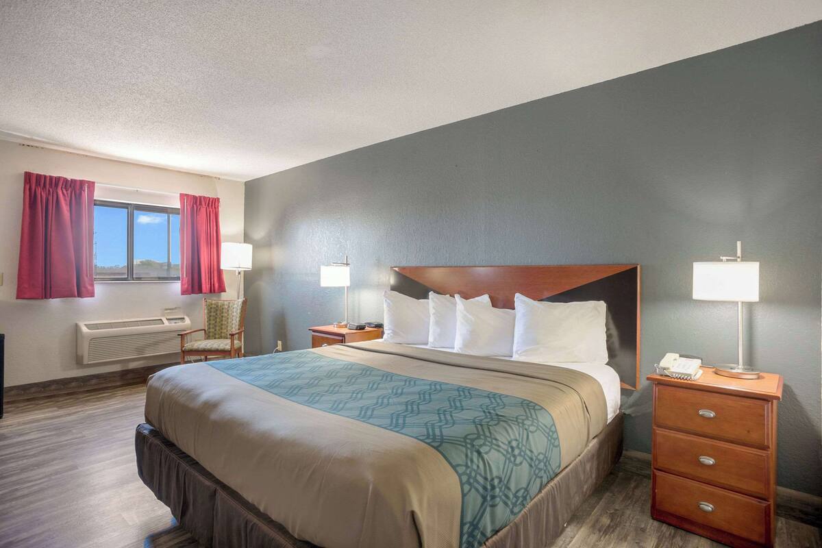 Habitación de hotel con una cama grande, cabecera de madera, dos mesitas de noche con lámparas, una silla y una ventana con cortinas.