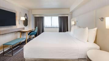 A hotel room with a large bed, a desk with a chair, a bench, a flat-screen TV, and a window with curtains.
