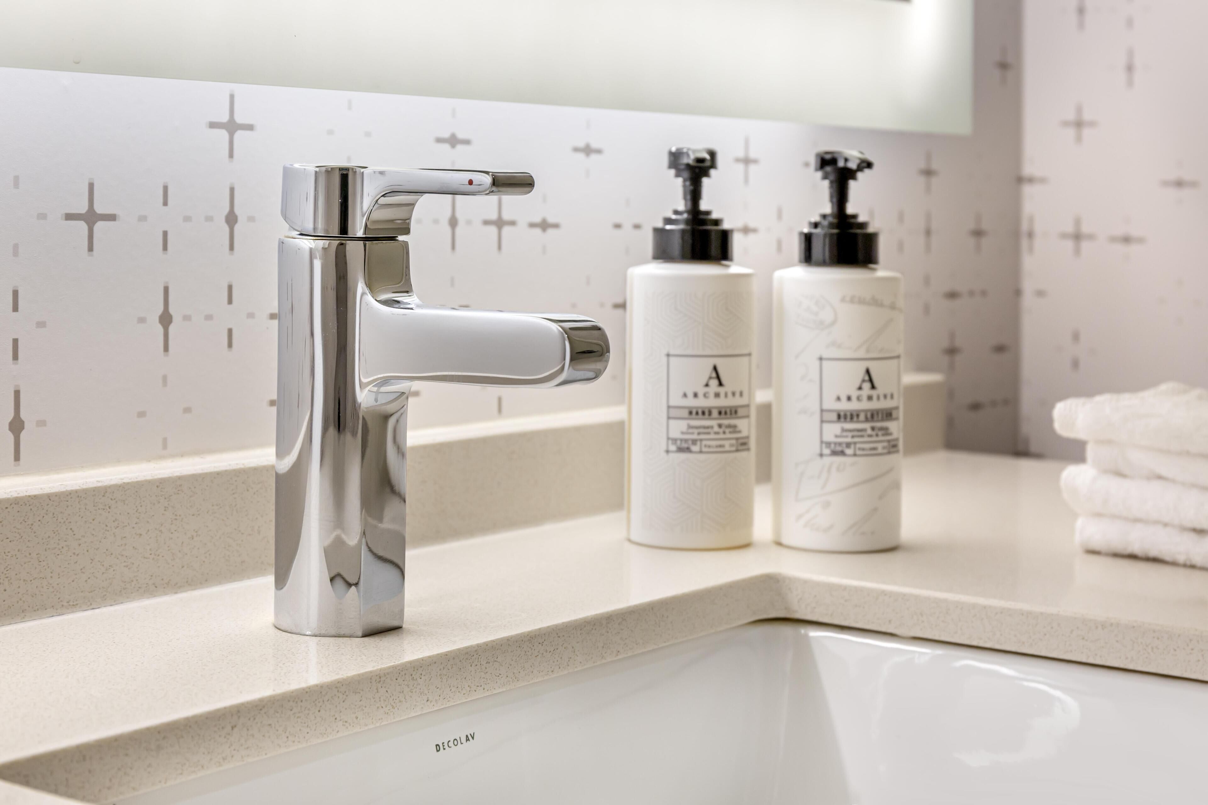A bathroom with a modern faucet, two bottles of hand soap, and a stack of white towels.