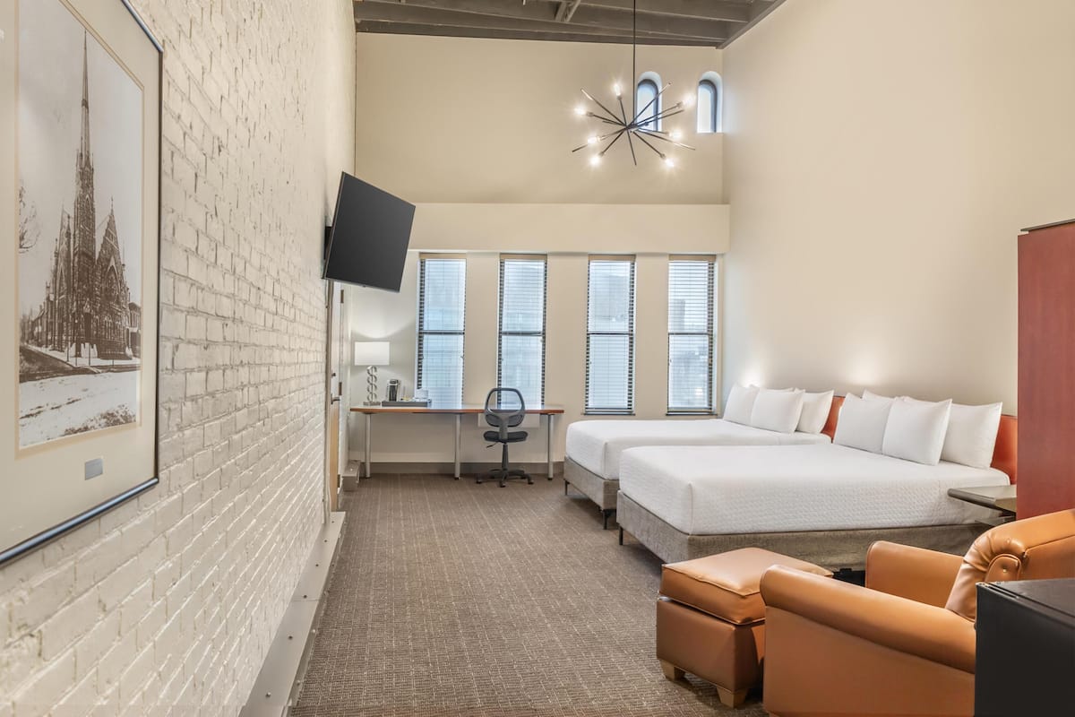 Quarto de hotel com parede de tijolos, uma cama, uma escrivaninha, uma cadeira, uma televisão e uma janela grande.