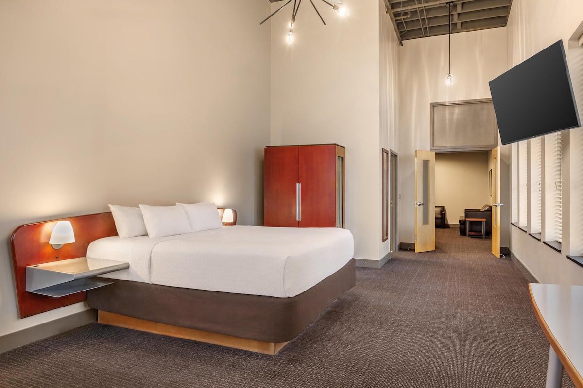 Quarto de hotel com cama, mesa de cabeceira, luminária, guarda-roupa, televisão e vista para o quarto vizinho.