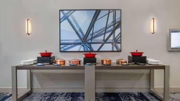 Una mesa de buffet con varias ollas de cobre y tapas rojas, frente a una pared con un cuadro abstracto grande y dos luces fijadas en la pared.