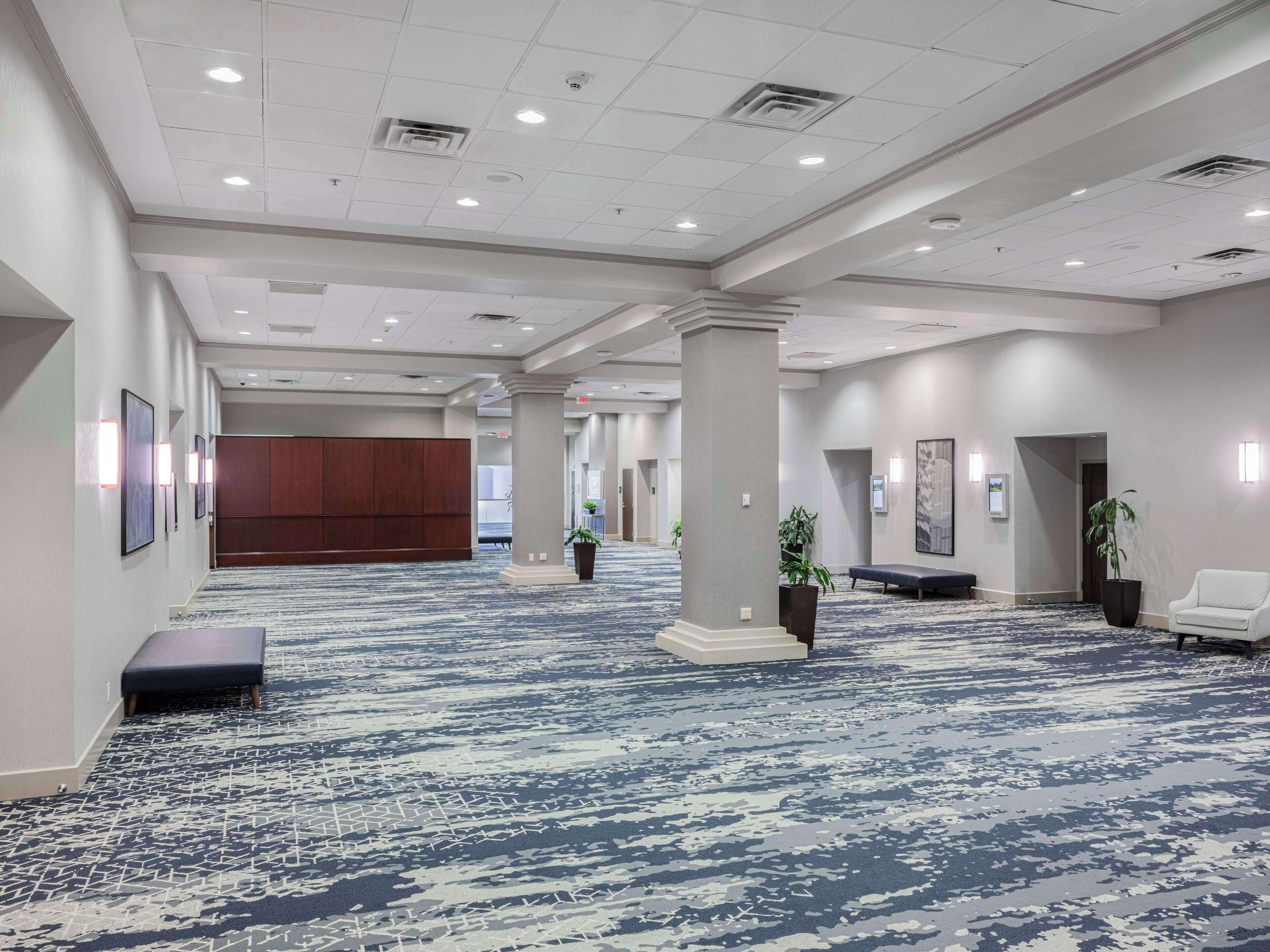 A spacious hotel lobby with a patterned carpet, large pillars, and a wooden panel wall.