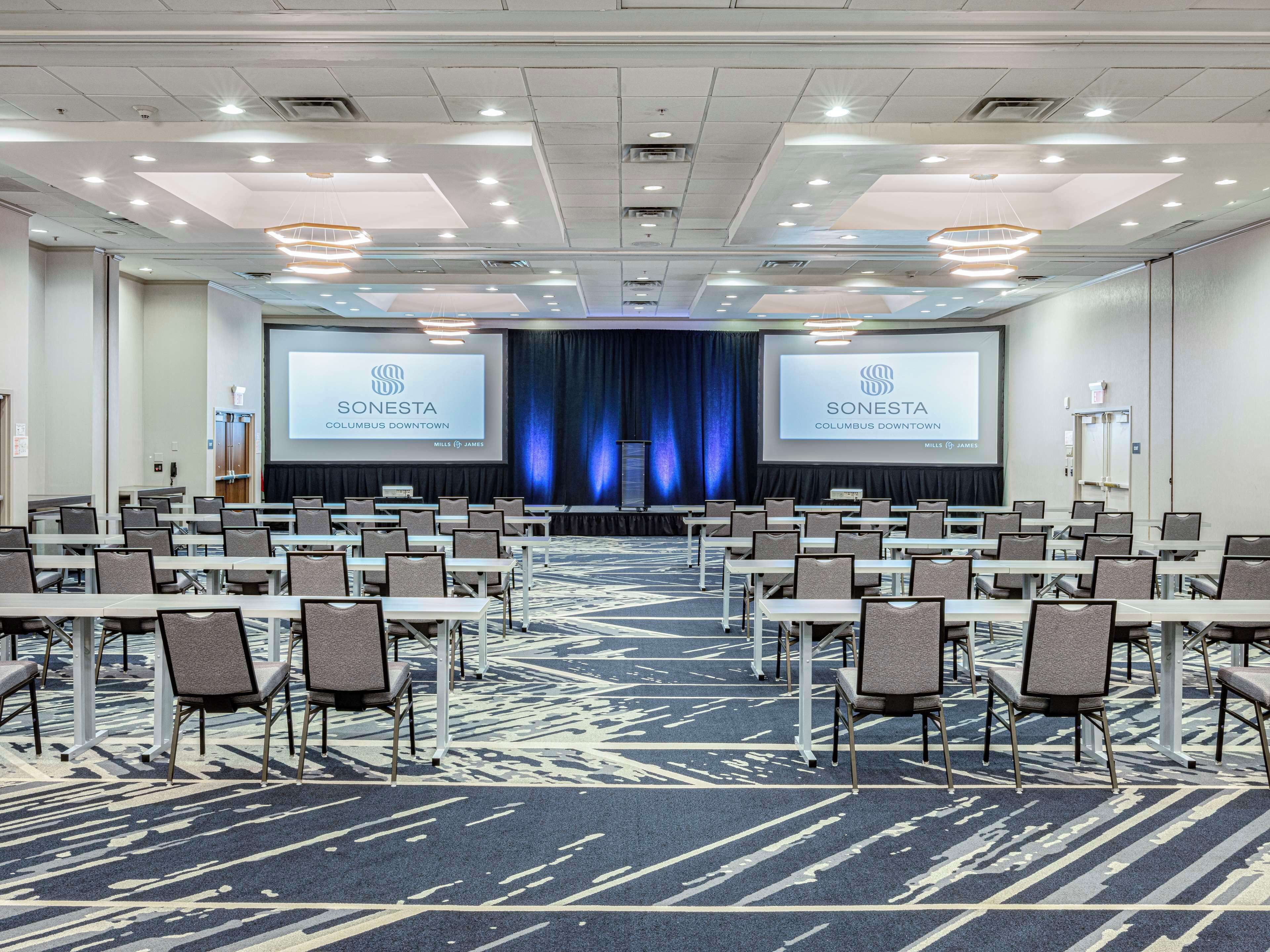 A conference room with rows of tables and chairs, a large screen displaying the Sonesta Columbus Downtown logo, and a patterned carpet.