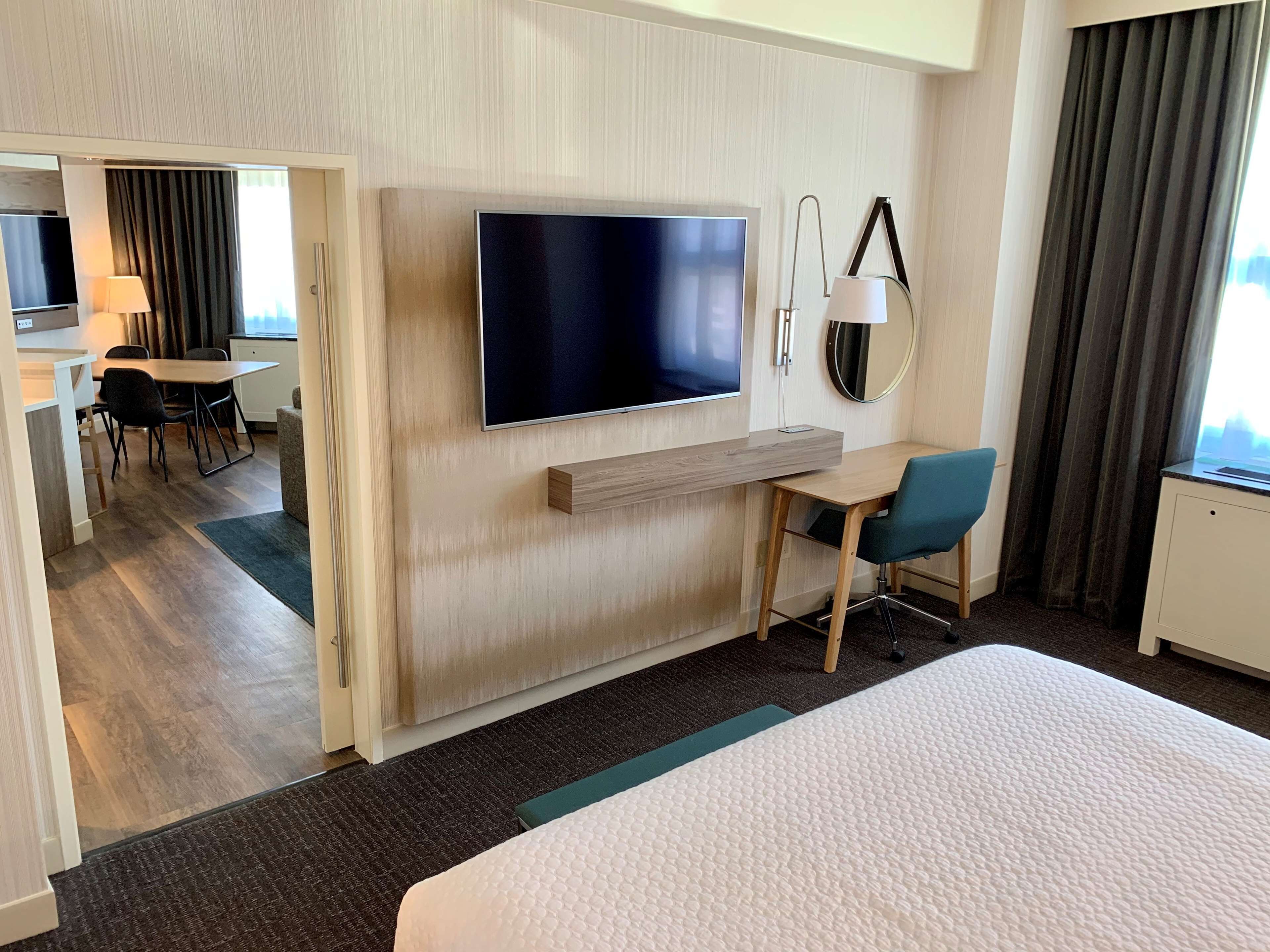 Ein Hotelzimmer mit einem großen Bett, einem Schreibtisch mit Stuhl, einem an der Wand befestigten Fernseher und Blick auf die Stadt durch das Fenster.