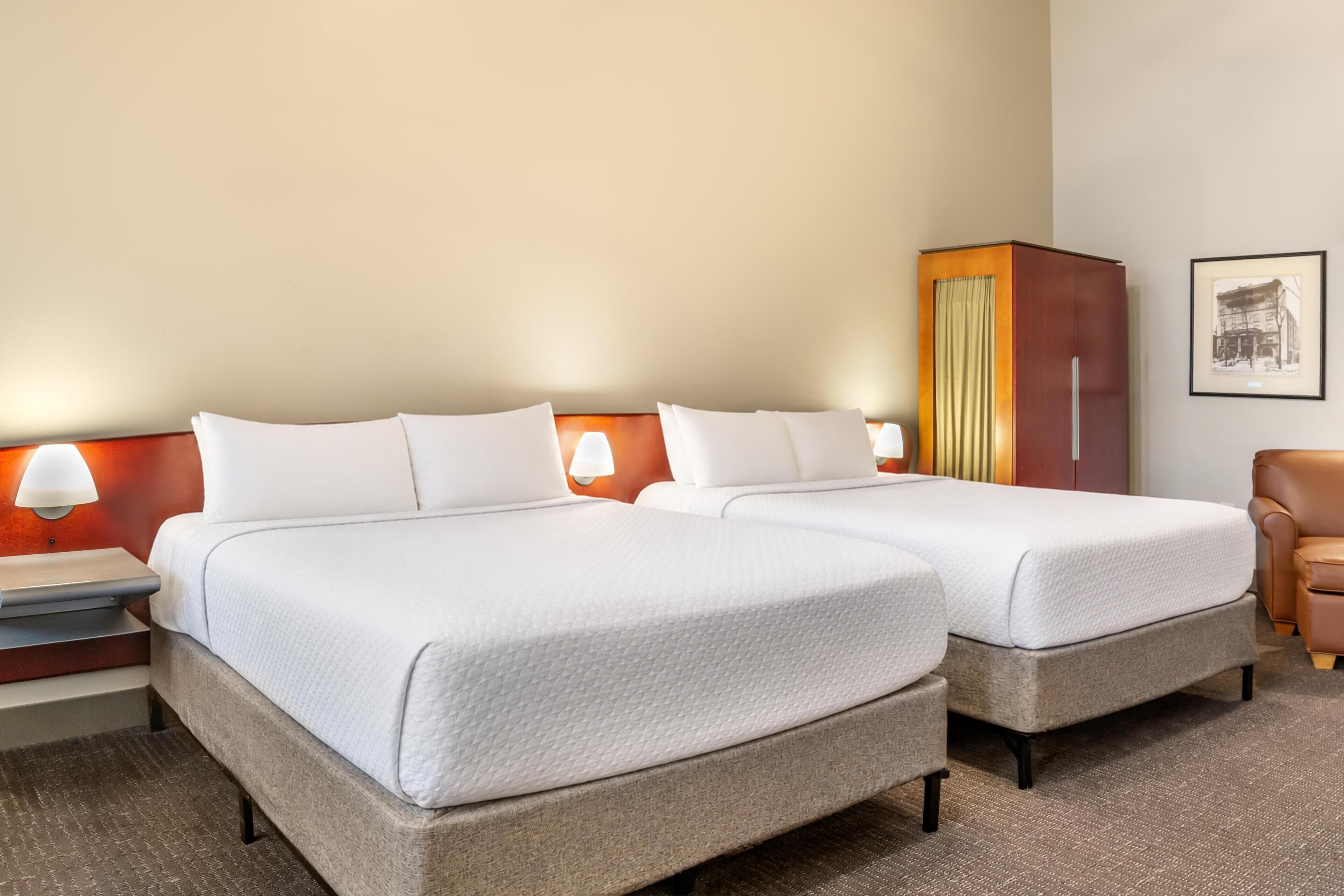 A hotel room with two beds, a nightstand, a lamp, a wardrobe, and a framed picture on the wall.