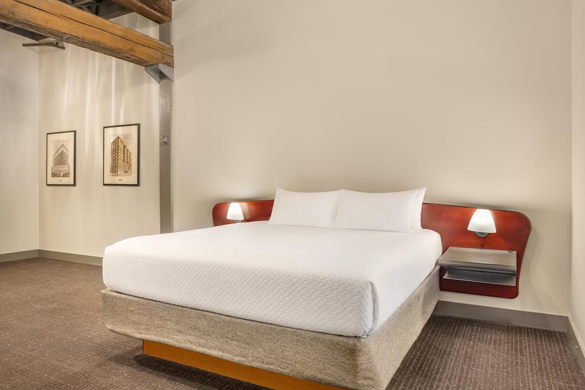 Cama bem arrumada com cabeceira de madeira, mesa de cabeceira com abajur e duas fotos emolduradas na parede.