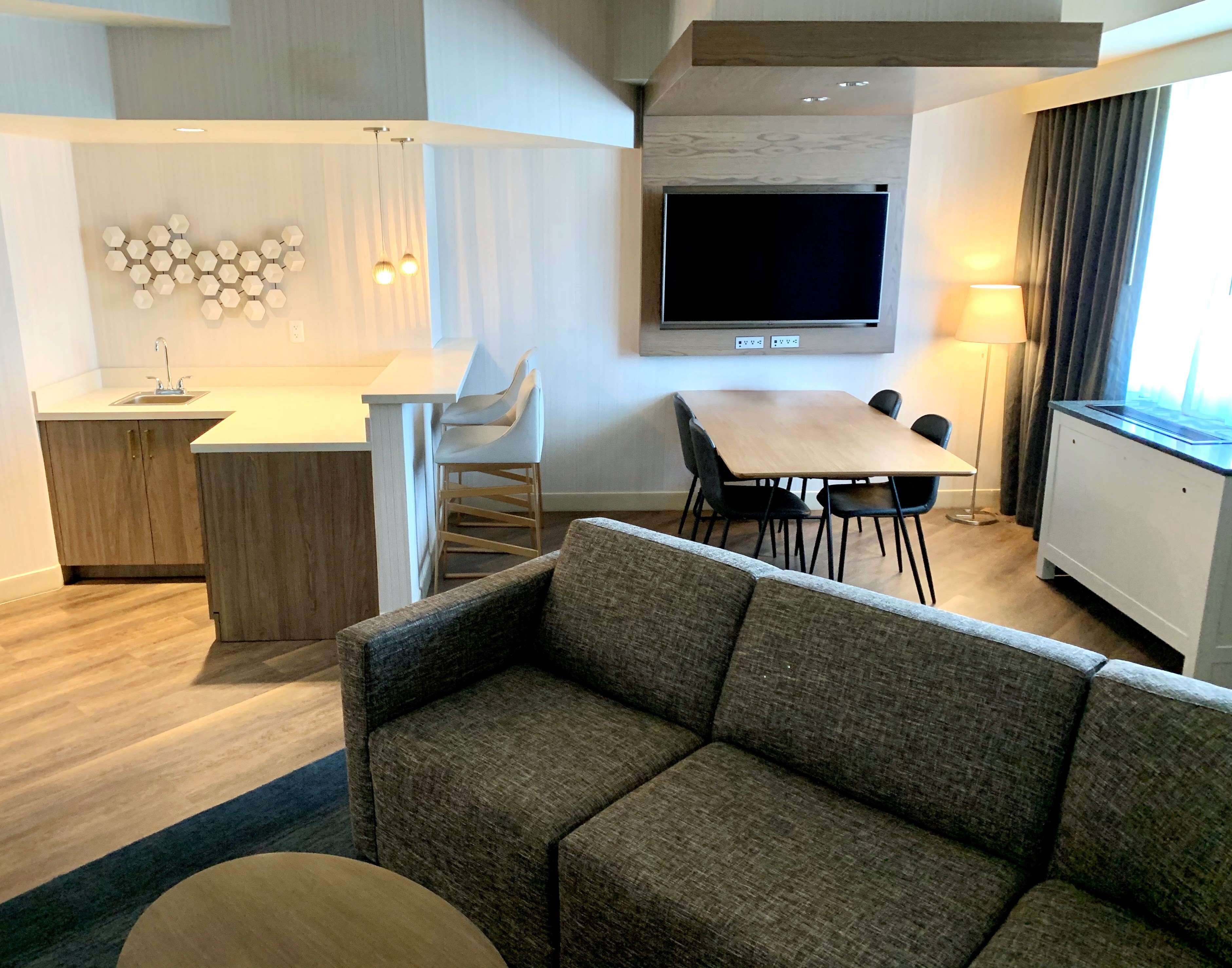 Ein modernes Wohnzimmer mit einer Couch, einem Esstisch mit Stühlen, einem an der Wand befestigten Fernseher und einer Küchenzeile mit Spüle und Schränken.