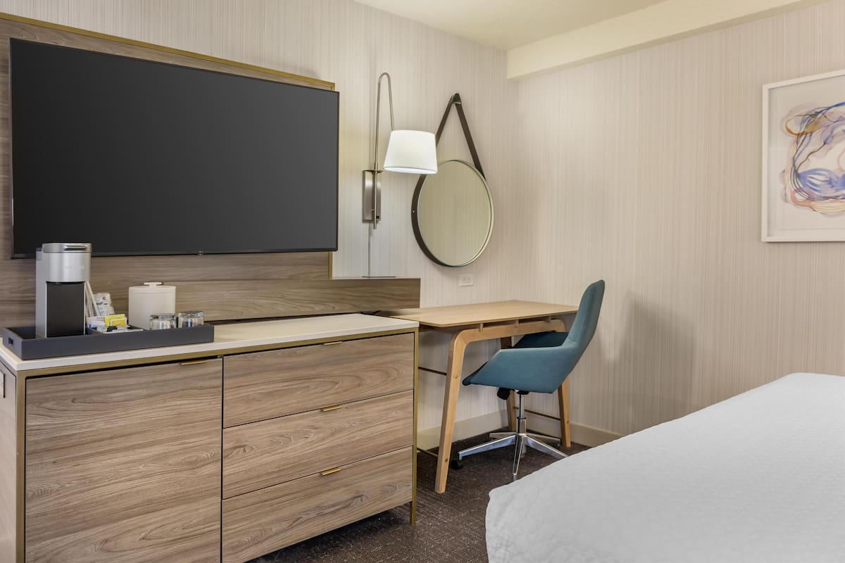Quarto de hotel com escrivaninha de madeira, uma cadeira, uma cama, televisão de tela plana, abajur, espelho redondo e uma pintura abstrata emoldurada.