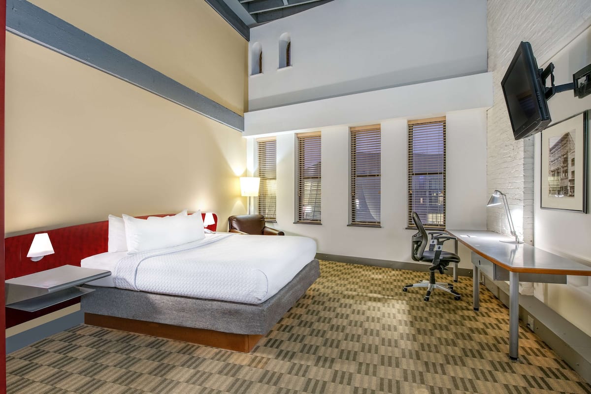 Quarto de hotel com cama, escrivaninha, cadeira, televisão e janelas.