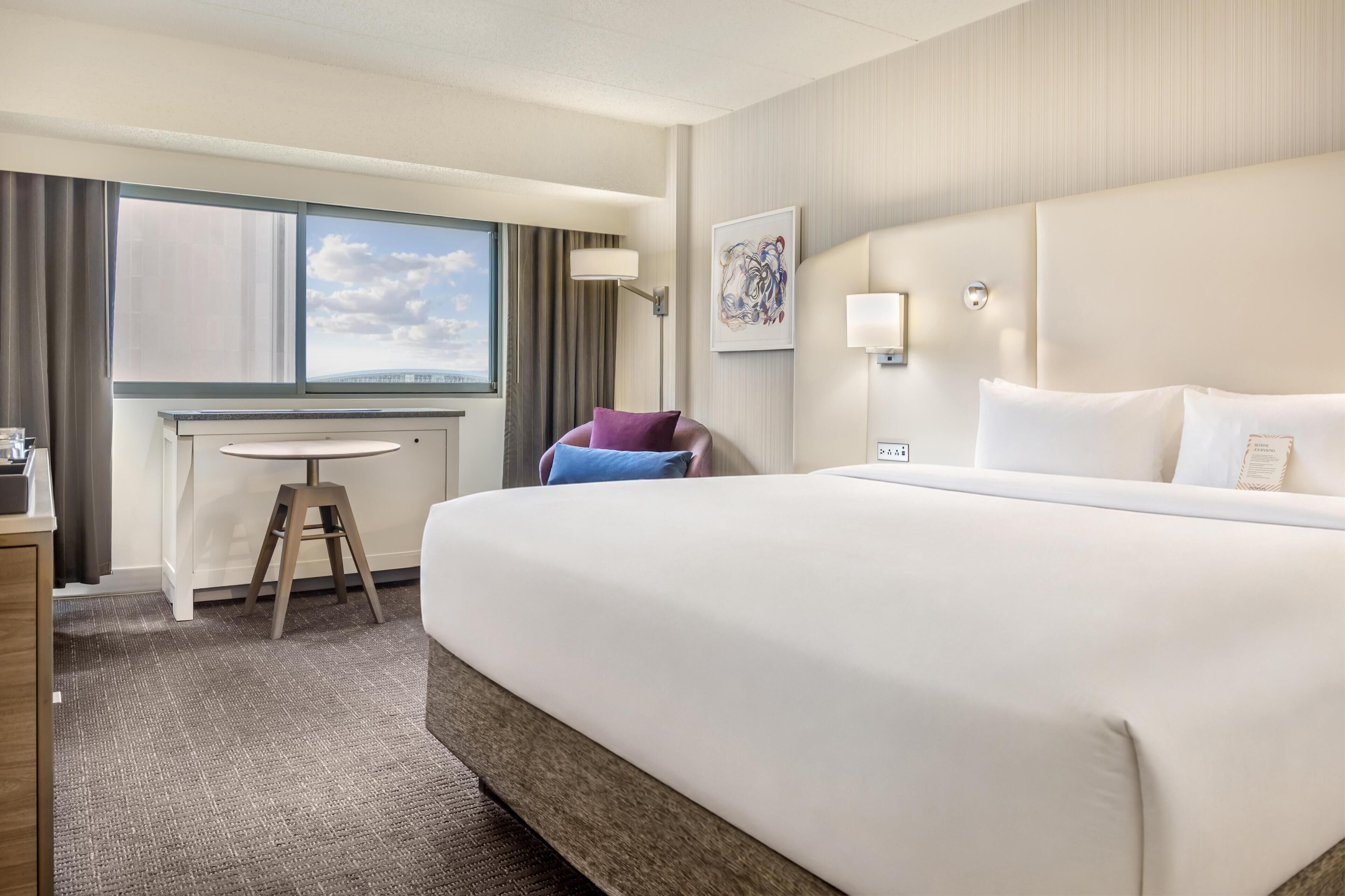 A hotel room with a large bed, a desk, a chair, a window with curtains, and a wall-mounted lamp.