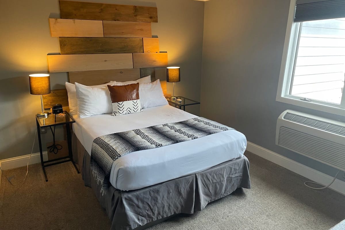A neatly made bed with a patterned bedspread and a grey skirted duvet. There are two bedside tables with lamps, a wooden headboard, and a window with blinds.