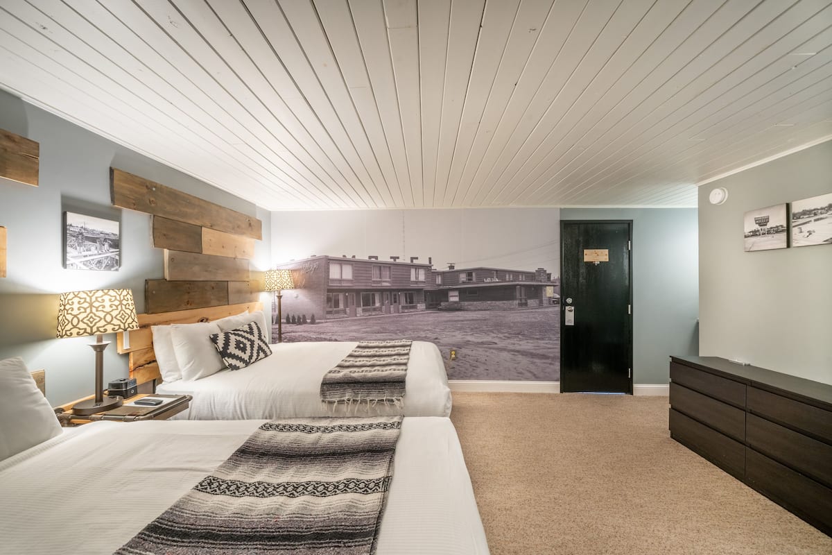 A hotel room with two beds, a wooden headboard, a wall mural, a dark wooden dresser, and a carpeted floor.