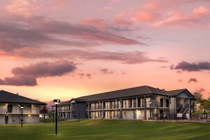 A modern hotel with multiple floors and balconies, featuring a well-maintained lawn and a sunset sky.