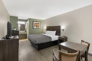 A hotel room with a bed, bedside table, chair, desk, television, and a painting on the wall.