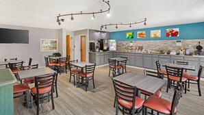 Un restaurante con varias mesas y sillas, una zona de cocina e imágenes de comida en una pared.