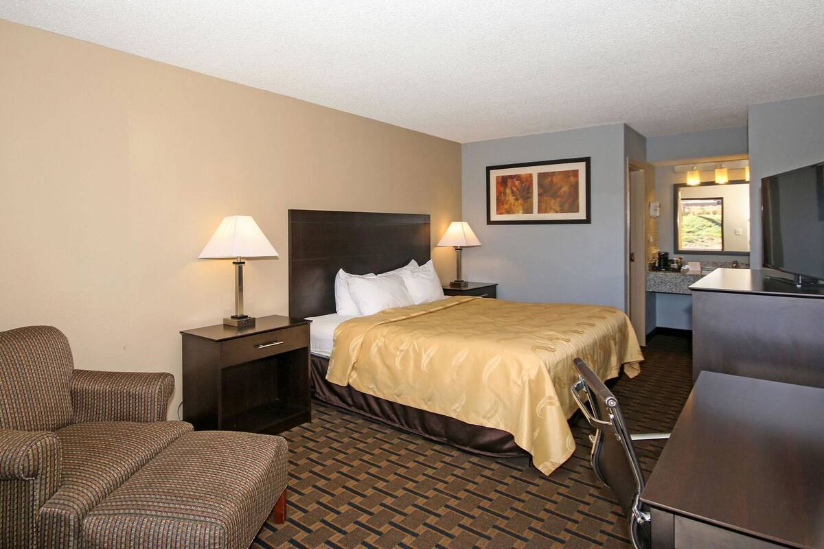 A hotel room with a bed, two bedside tables with lamps, a chair, a desk, and a kitchenette area.