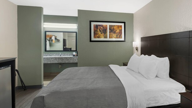 Habitación de hotel con cama, mesita de noche, espejo, un cuadro abstracto enmarcado y un baño visible a través de una puerta abierta.
