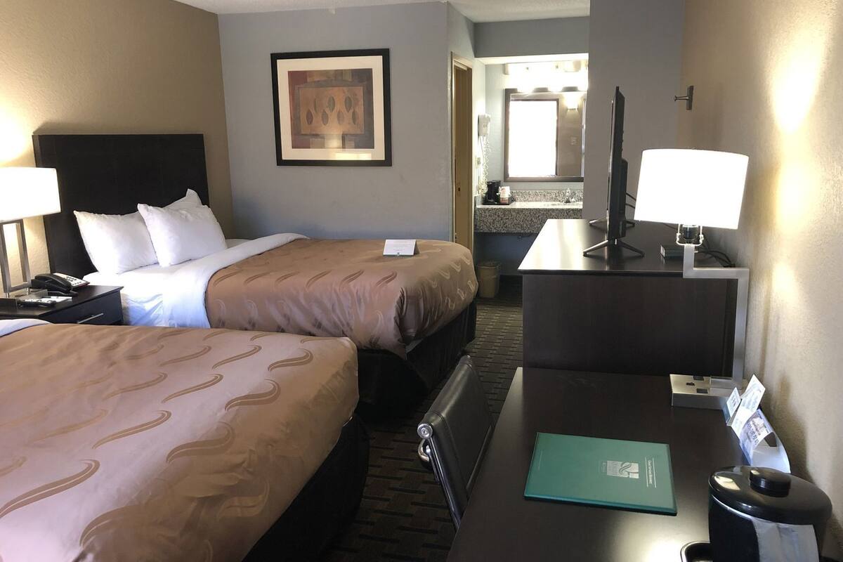 A hotel room with two beds, a desk with a lamp, a TV, a bathroom visible through an open door, and a framed picture on the wall.