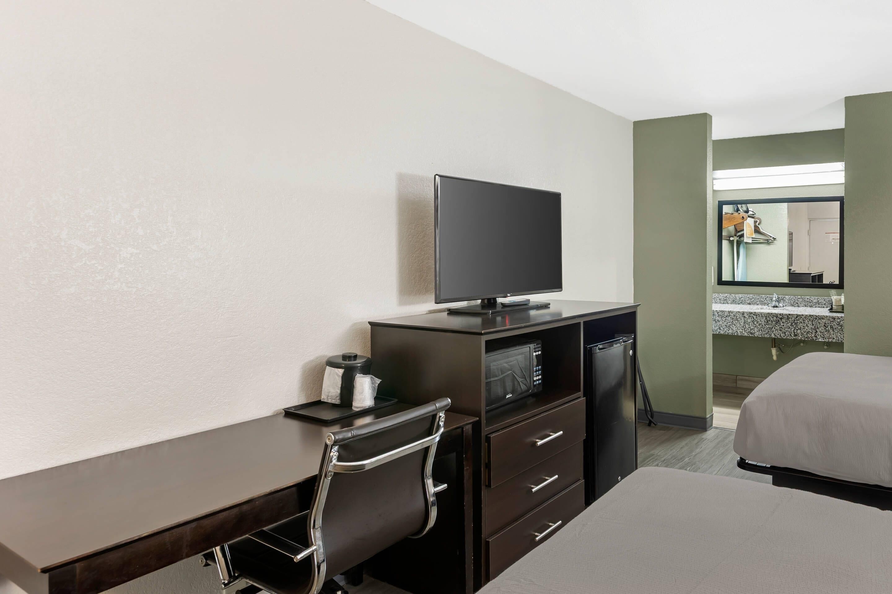 Una habitación de hotel con cama, escritorio, televisor, microondas y baño con lavamanos y espejo.