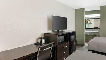 Una habitación de hotel con cama, escritorio, televisor, microondas y baño con lavamanos y espejo.