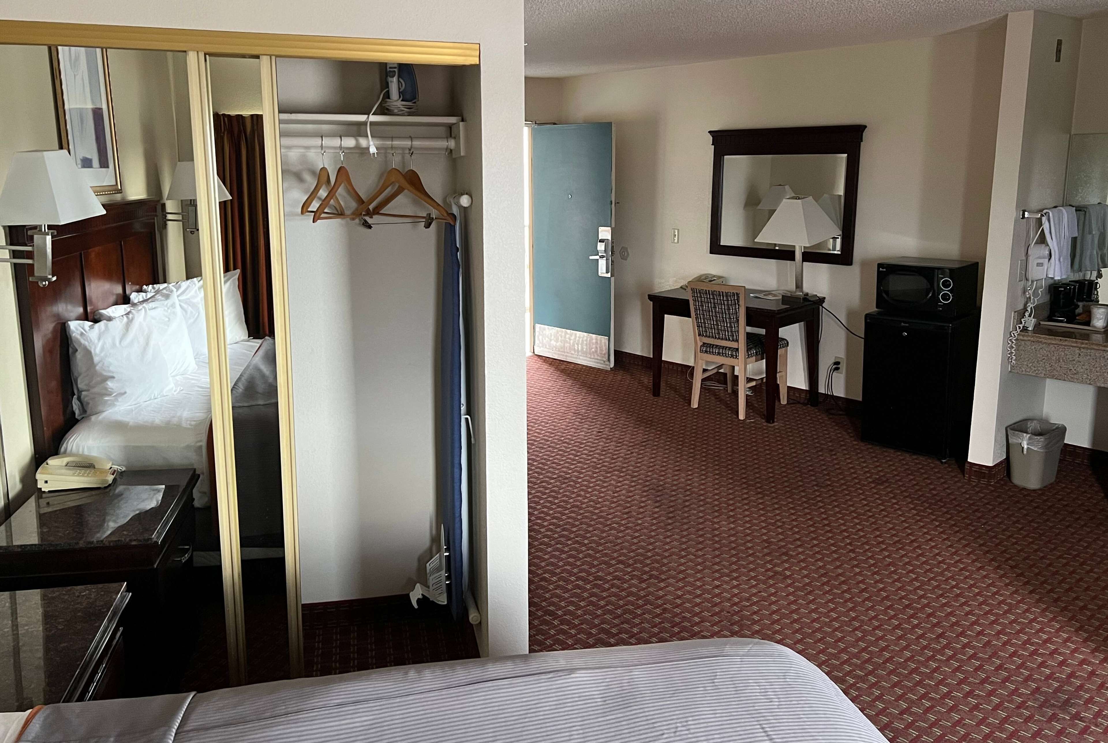 A hotel room with a bed, bedside table, lamp, and a mirror. There is a desk with a chair and a microwave.