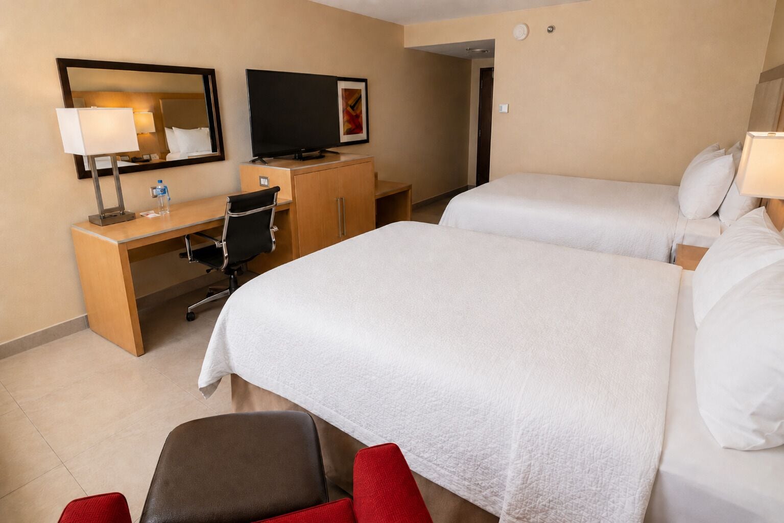 Una habitación de hotel con dos camas, un escritorio con una silla, un televisor y un espejo.