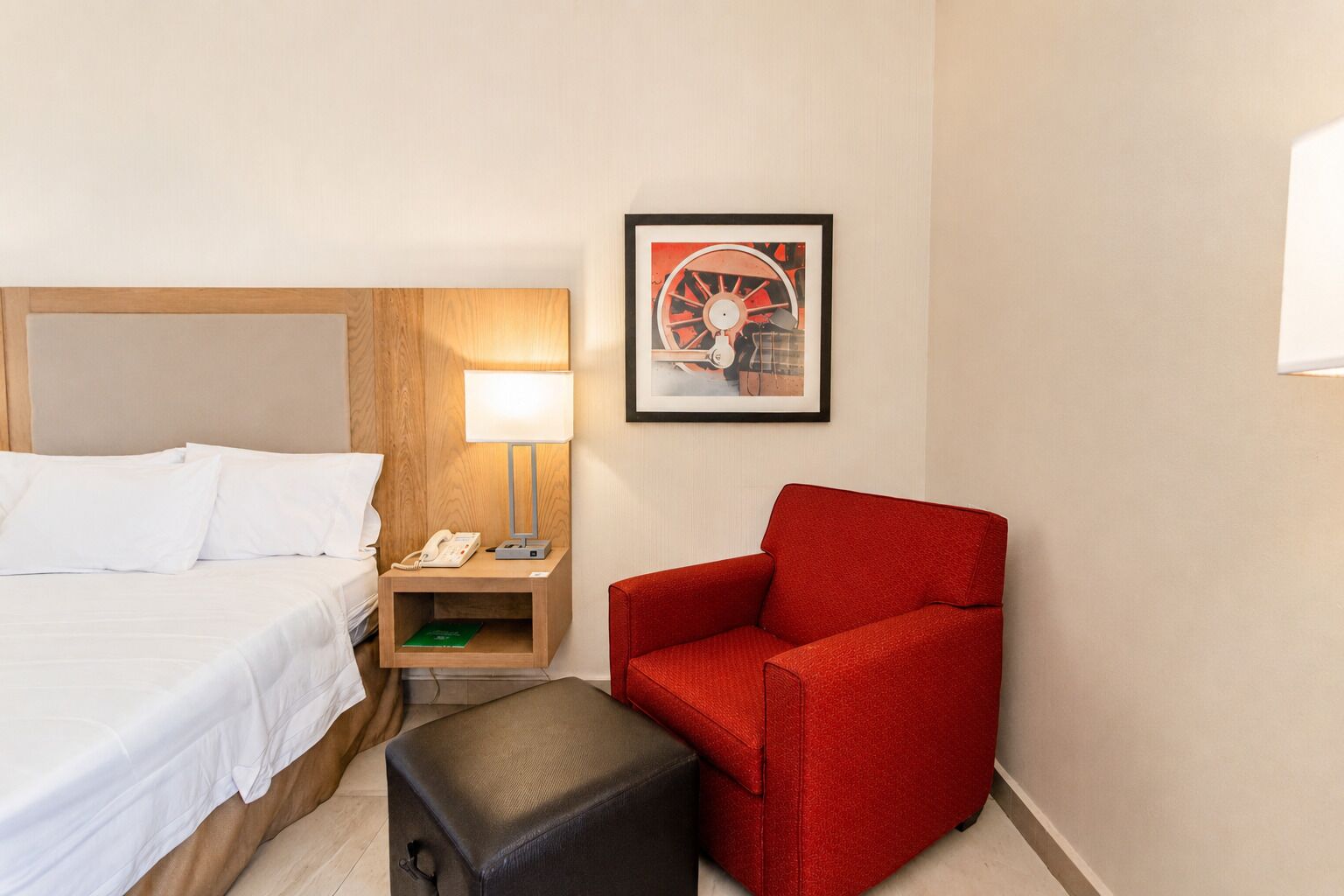 Una habitación de hotel con una cama, una silla roja, una mesita de noche con una lámpara y un cuadro en la pared.