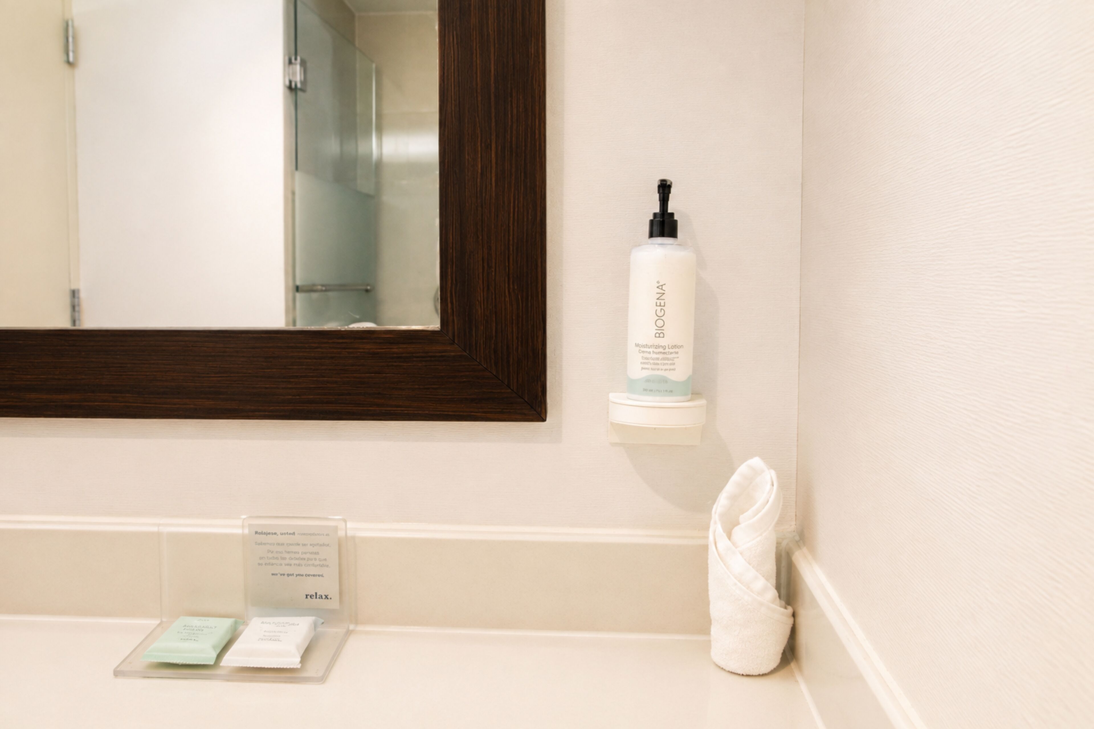 Une salle de bain avec un miroir, une bouteille de lotion hydratante Biogena et une serviette.