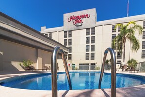 A swimming pool with metal ladders in front, a Hampton Inn sign, and a building with multiple windows in the background.