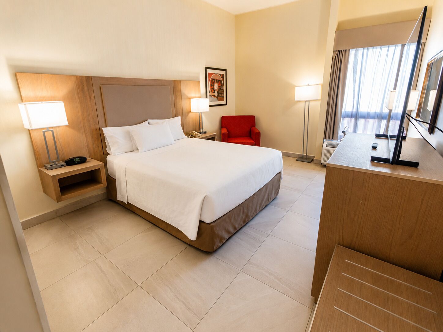 Una habitación de hotel con cama, mesitas de noche, una silla, un escritorio y un ventanal grande.