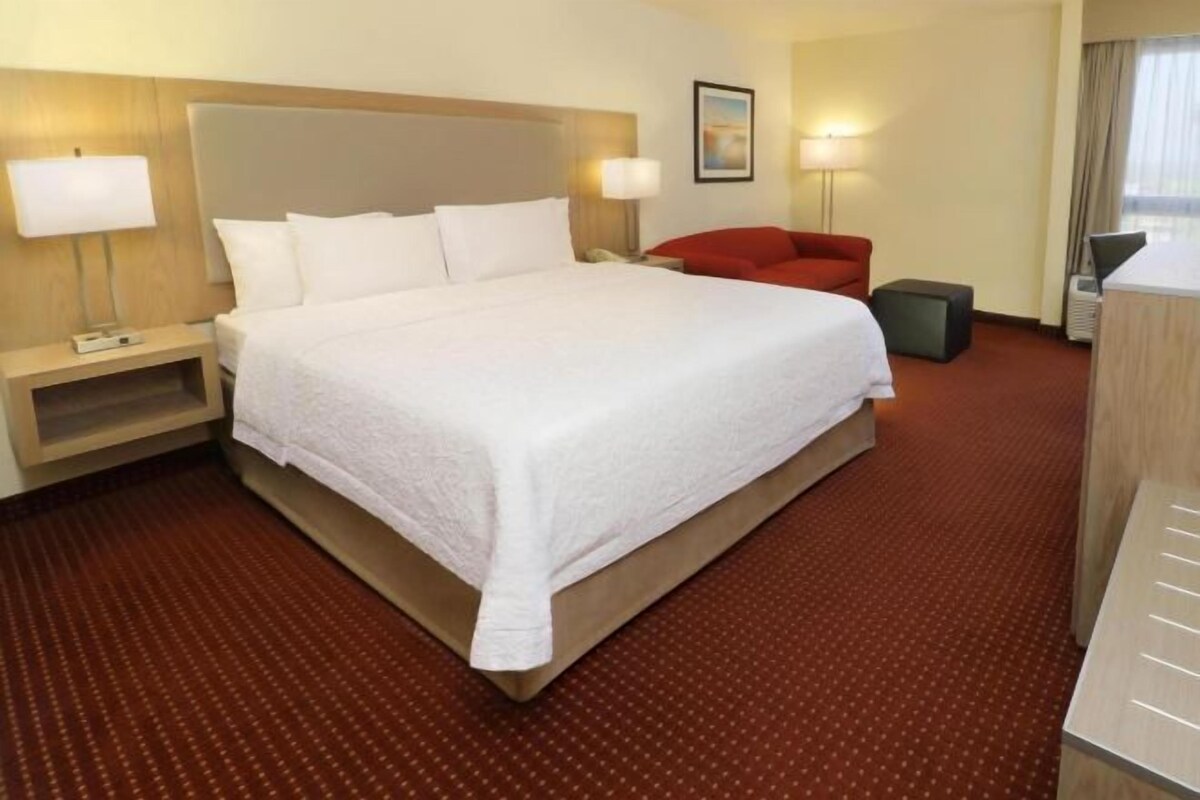 A hotel room with a large bed, bedside lamps, a red chair, and a desk with a lamp.