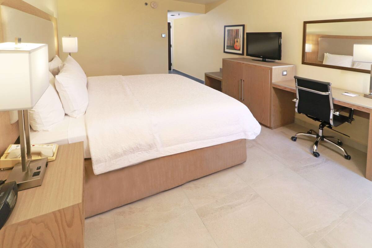 A hotel room with a large bed, a desk with a computer, a chair, a TV, and a mirror.
