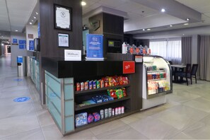 A hotel reception area with a self-service beverage machine, a refrigerated display case, and a seating area with a view of the windows. - Hampton Inn by Hilton Monterrey-Airport (Apodaca)