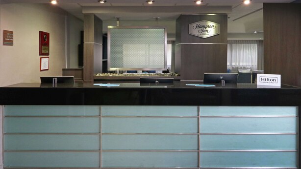 A reception desk with a Hampton Inn sign, a computer, and a glass partition. - Hampton Inn by Hilton Monterrey-Airport (Apodaca)