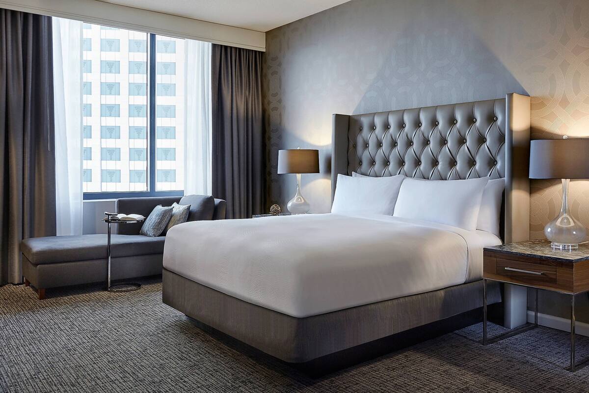 Une chambre d’hôtel moderne avec un grand lit, un canapé, deux tables de chevet et une vue sur un immeuble par la fenêtre.