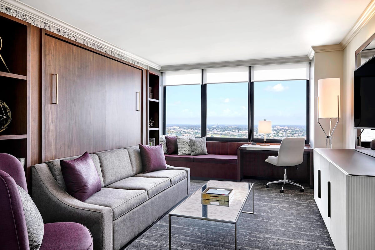 Un salon moderne comprenant un canapé, des fauteuils, une table basse et un bureau avec une lampe. De grandes fenêtres offrent une vue sur la ville.