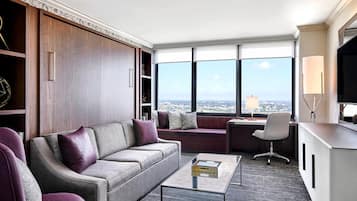 A modern living room with a sofa, armchairs, a coffee table, and a desk with a lamp. Large windows offer a city view.