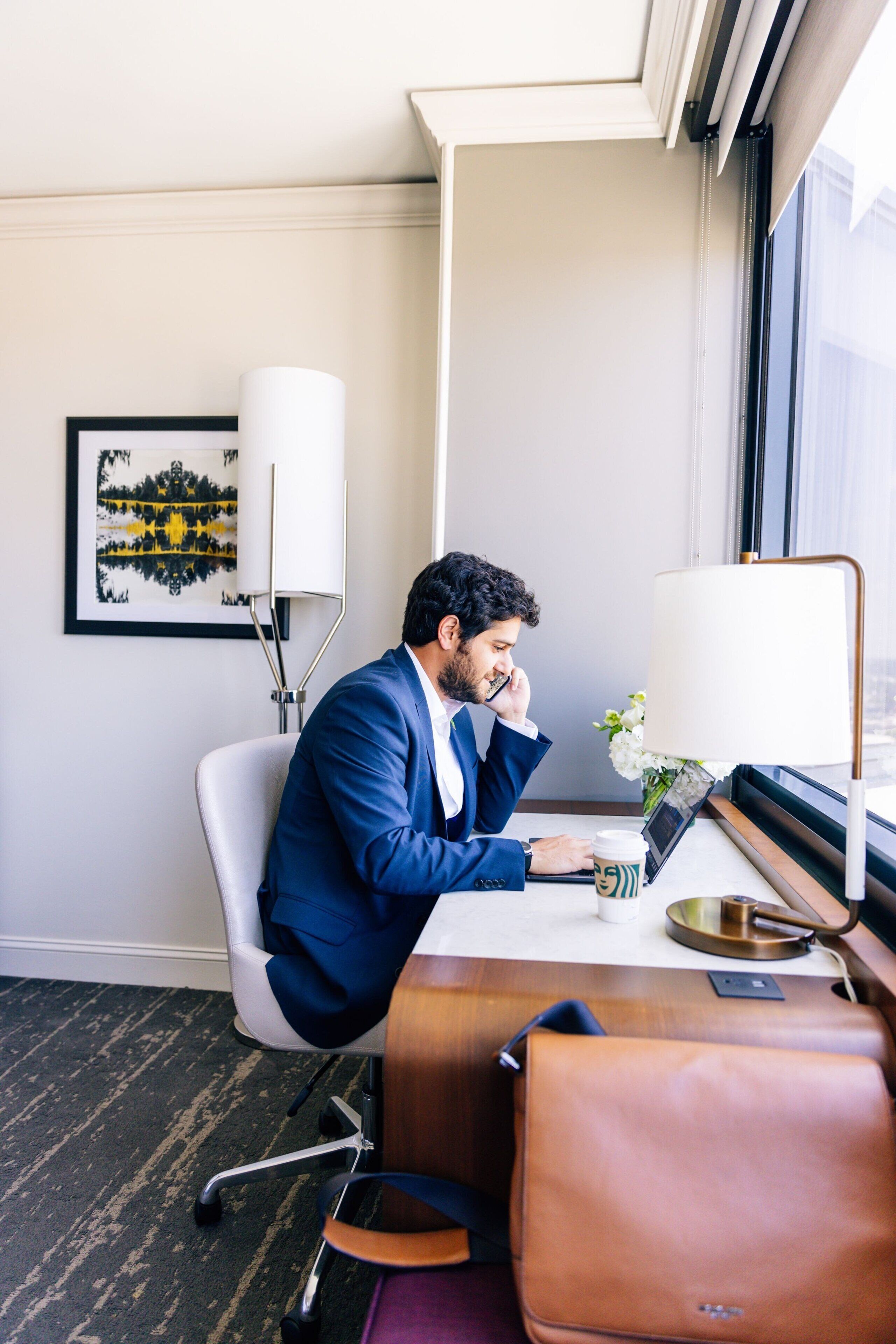 A man in a blue suit sitting at a desk in a hotel room with a laptop, a coffee cup, and a lamp.
