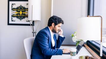 A man in a blue suit sitting at a desk in a hotel room with a laptop, a coffee cup, and a lamp.