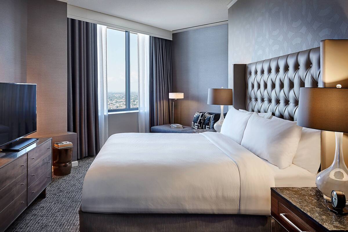 Une chambre d’hôtel moderne dotée d’un grand lit, d’une télévision à écran plat, d’une table de chevet avec une lampe et offrant une vue sur la ville grâce à de grandes fenêtres.