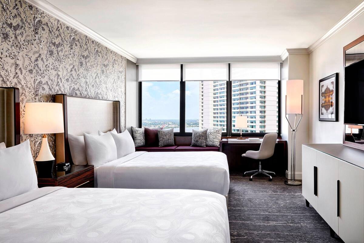 Une chambre d’hôtel avec deux lits, un bureau, une chaise, une télévision et une grande fenêtre donnant sur la ville.
