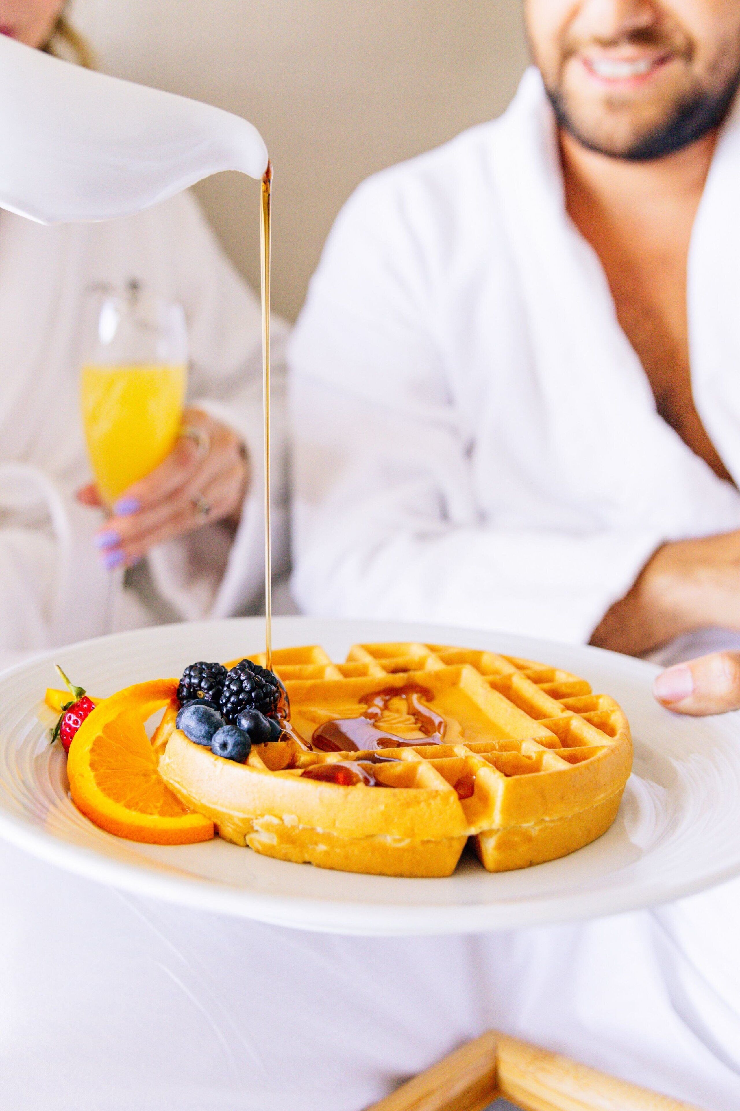 A breakfast plate with waffles, blueberries, orange slices, and a strawberry, with syrup being poured over it.