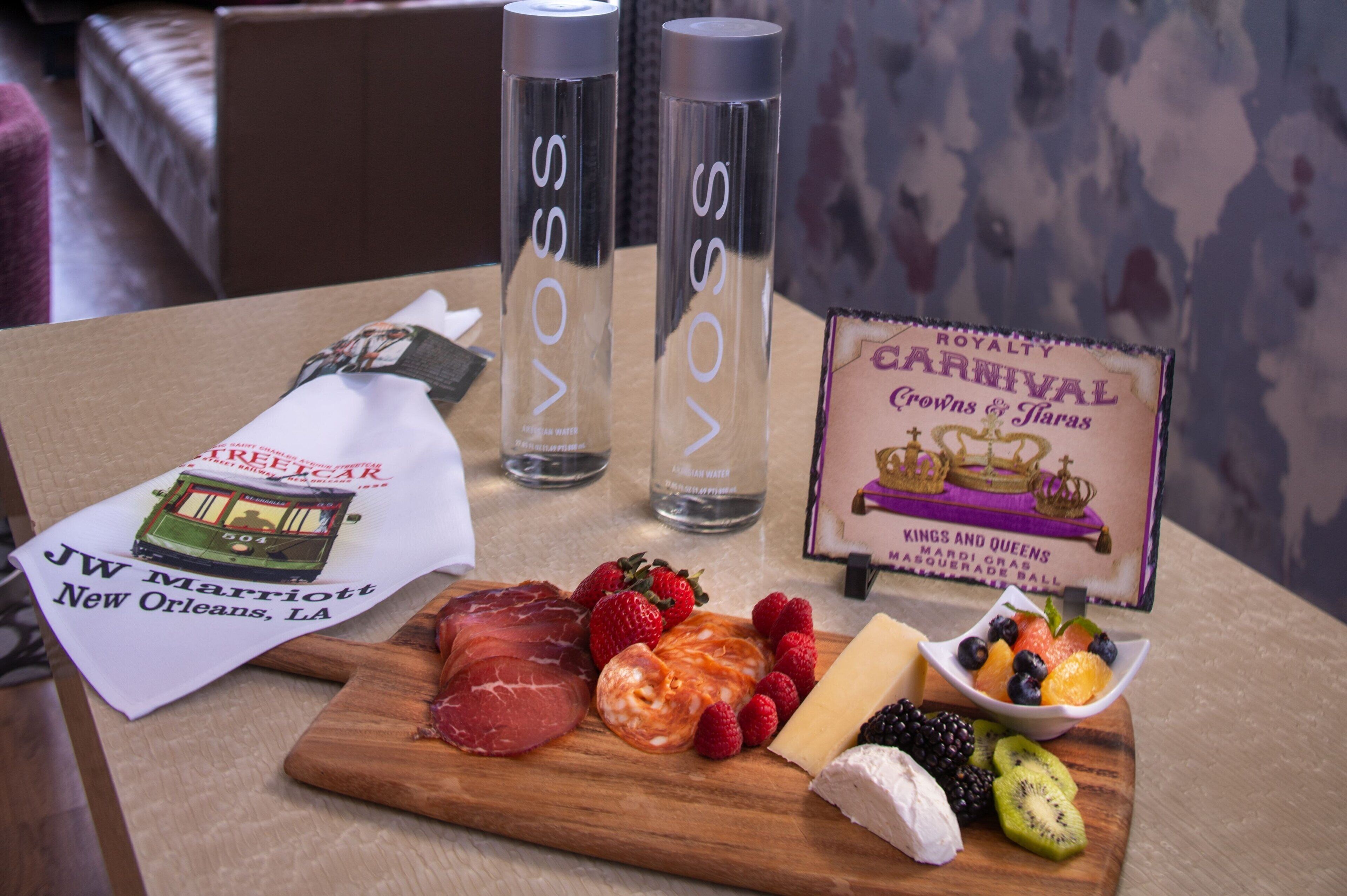 Uma mesa com tábua de madeira exibindo diversos queijos, frutas e carnes. Há duas garrafas transparentes com a etiqueta "VOSS" e uma placa para "ROYALTY CARNIVAL" com desenho de coroa e bola. Um folheto de hotel também está visível.