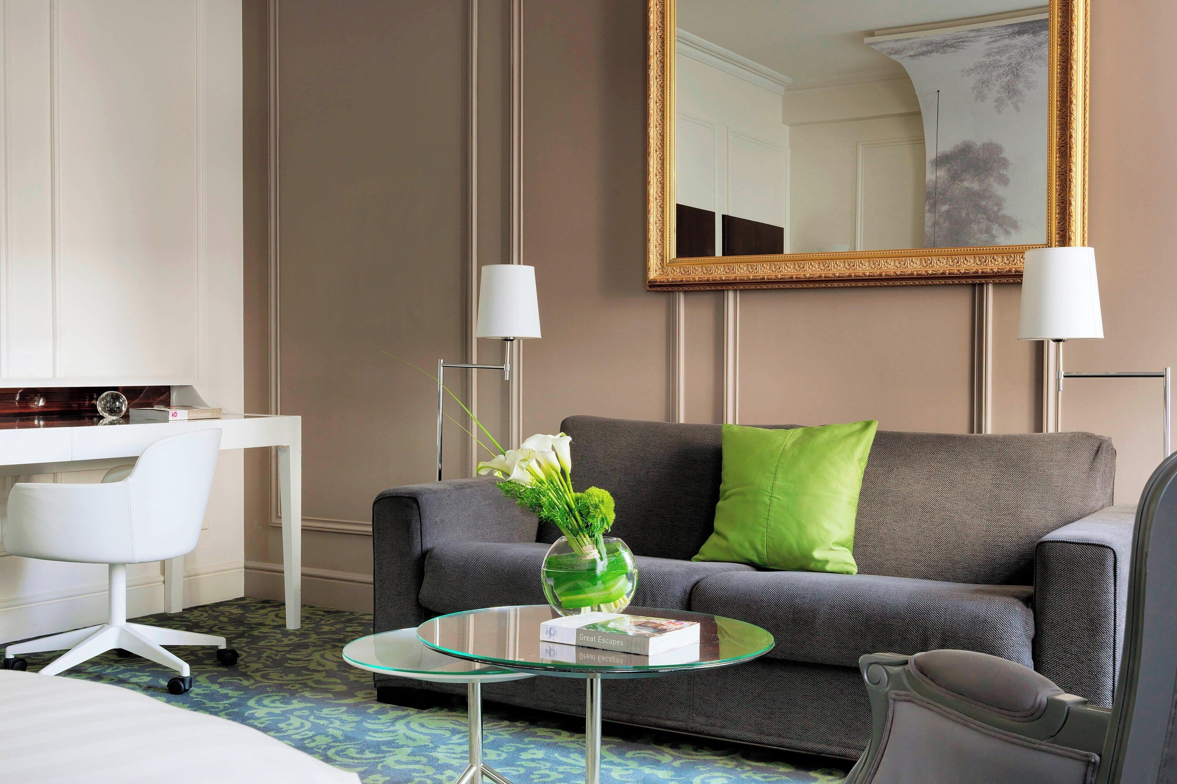 Ein Hotelzimmer mit grauem Sofa, weißem Schreibtisch, Glastisch, grünem Kissen und Spiegel mit goldener Rahmung.