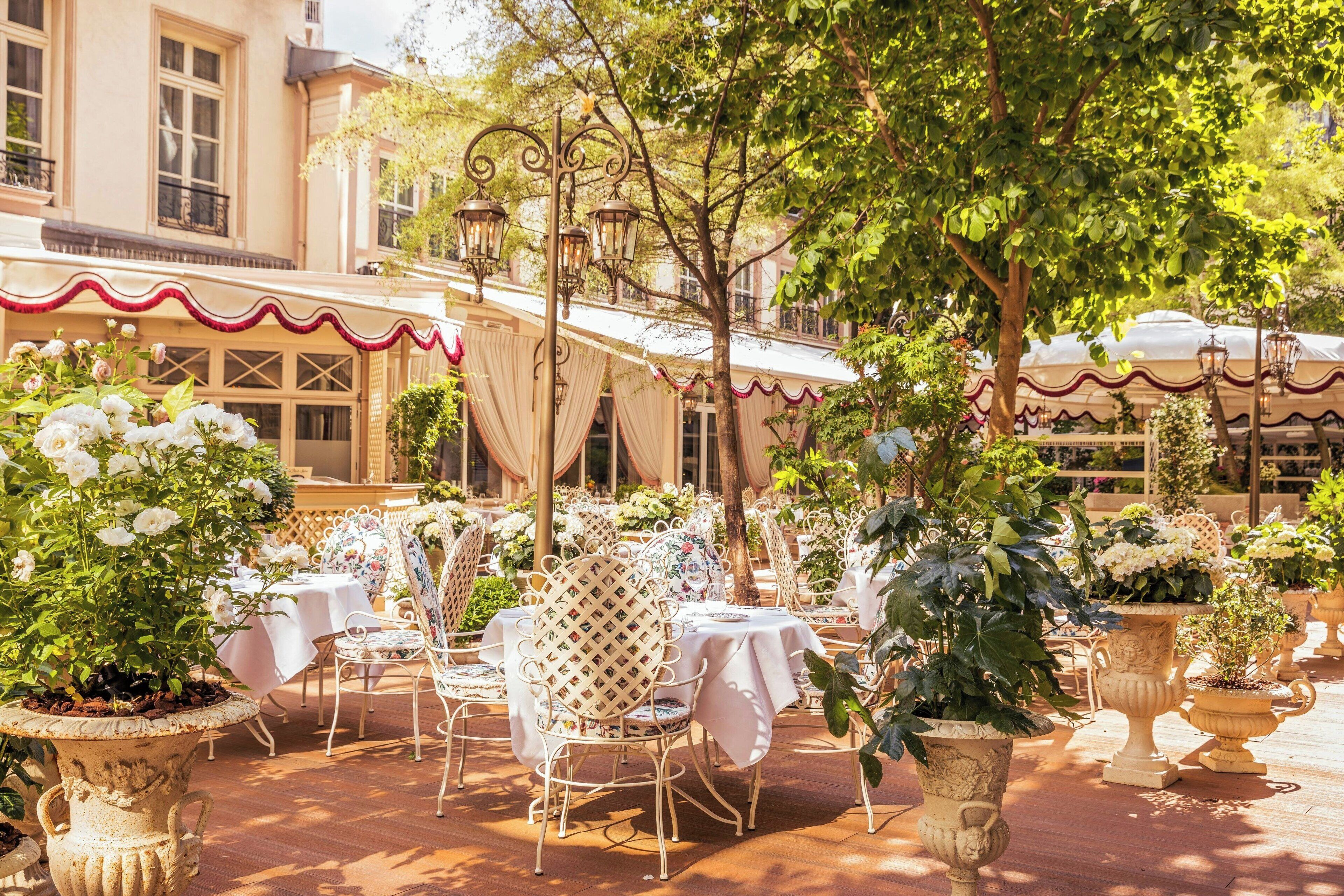 An outdoor dining area with tables and chairs, surrounded by potted plants and a building with a balcony.