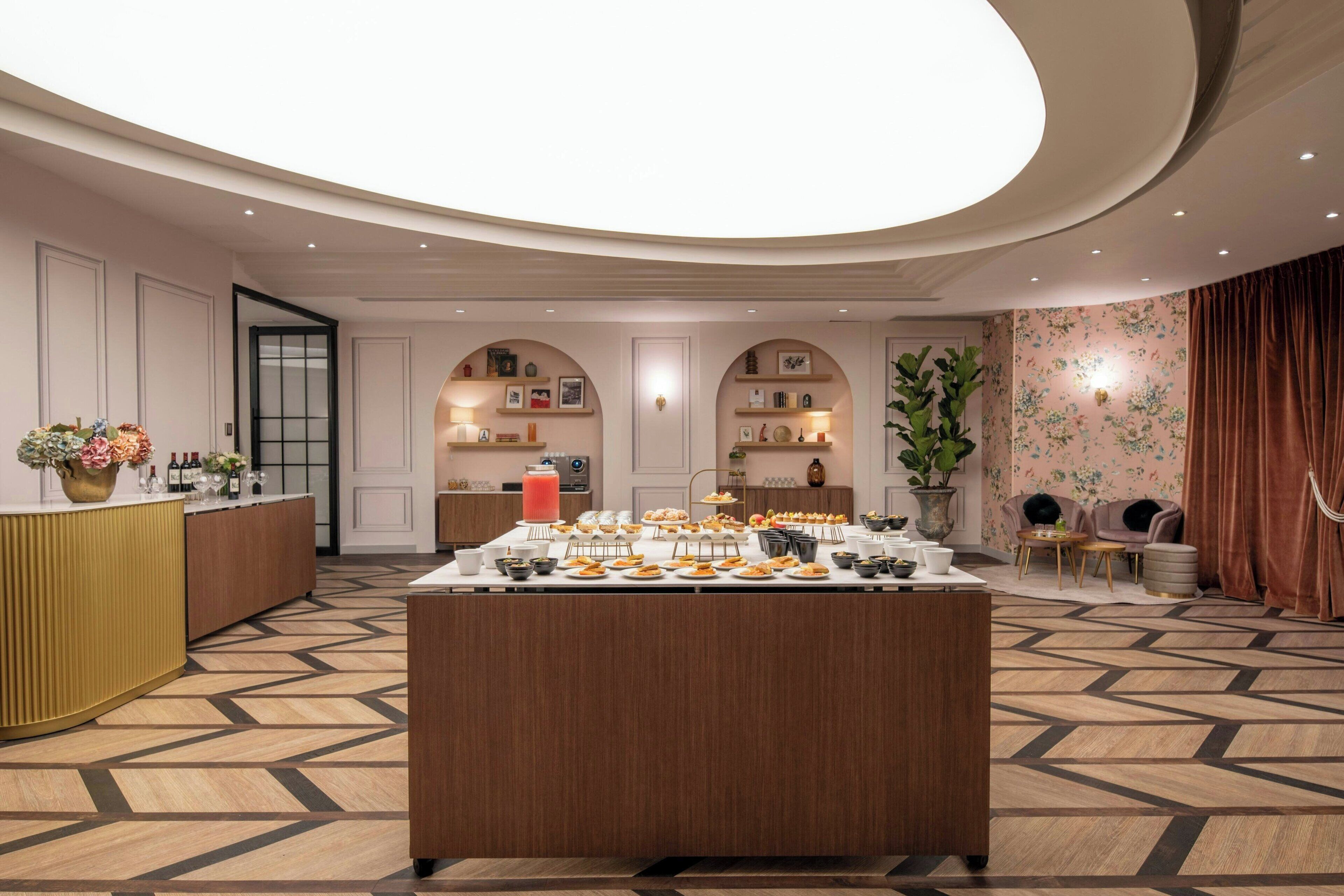 A hotel lobby with a central buffet table, a reception desk, and a seating area with a plant and curtains.