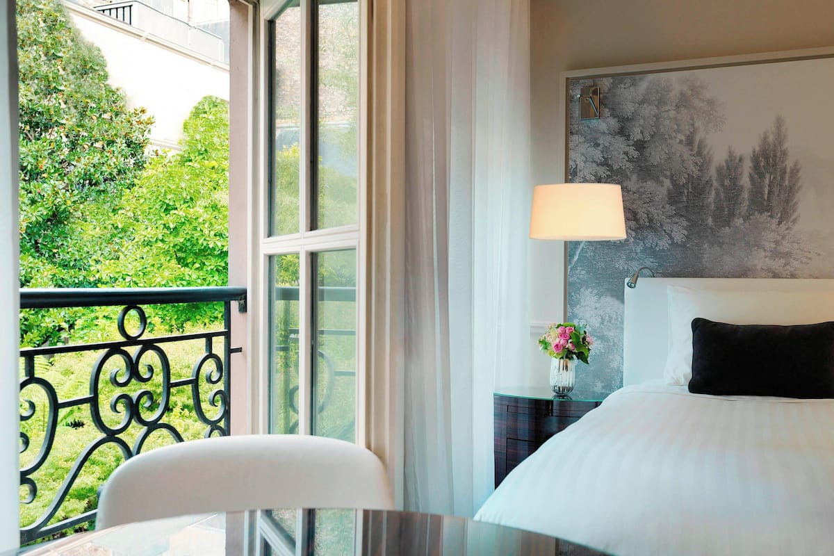 A hotel room with a bed, a nightstand, a lamp, a window with a view of greenery, and a painting on the wall.
