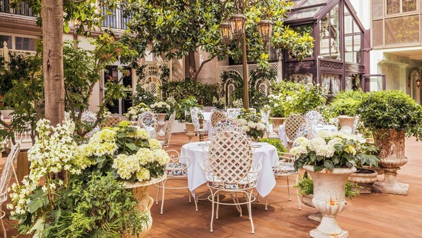 Un espace de restauration en plein air avec des tables et des chaises blanches, entouré d’une végétation luxuriante et de plantes en pot.